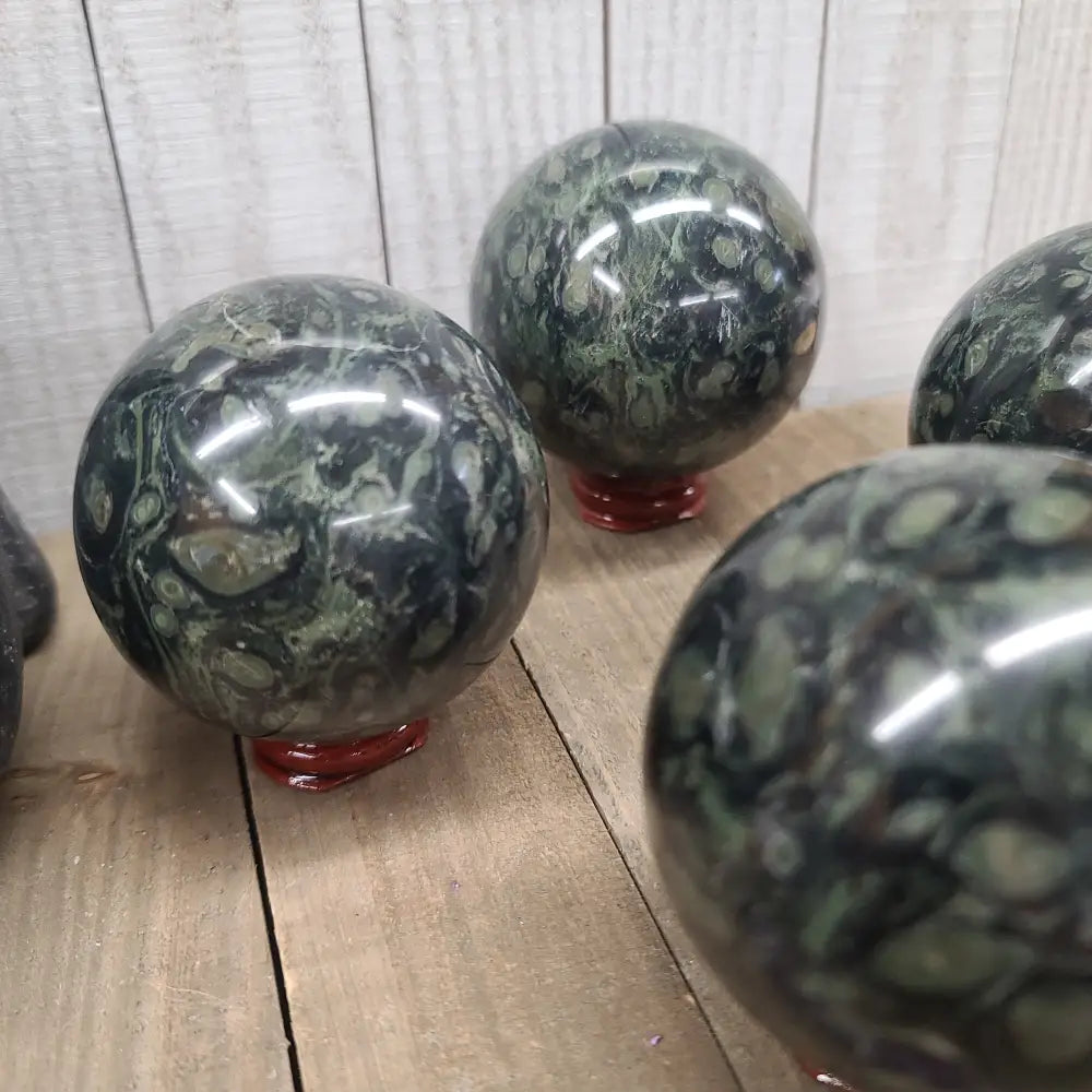 Hand-polished Kambaba Jasper sphere featuring deep green and black swirling orbicular patterns, displayed on a white background.