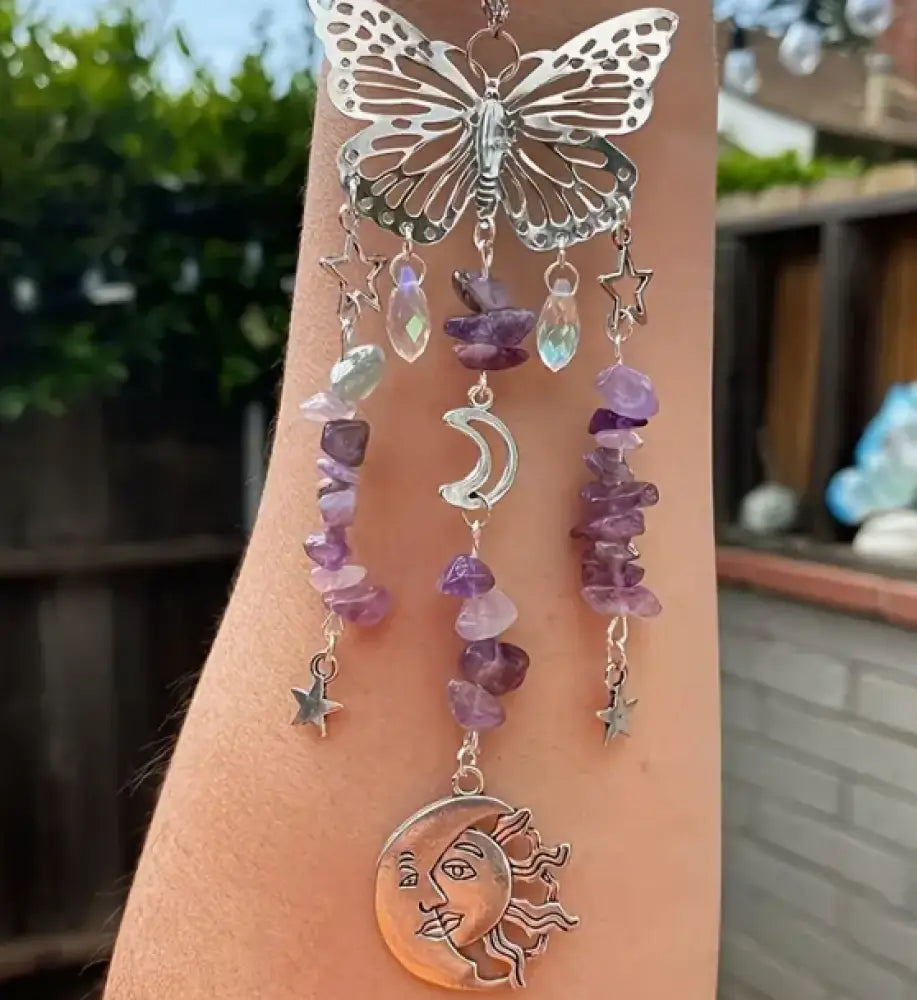 Silver butterfly and moon charm necklace with purple stones on a hand against a blurred outdoor background