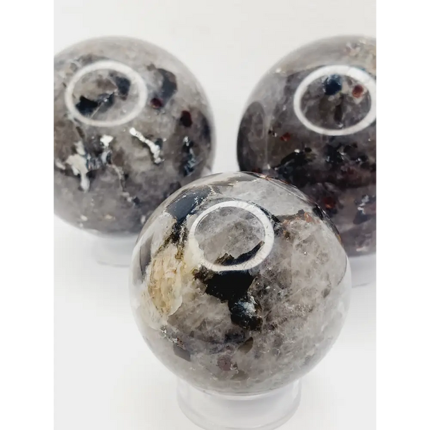 Quartz, garnet, tourmaline, and mica sphere, polished with clear, red, black, and shimmering inclusions, displayed on a white background.
