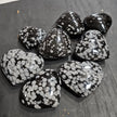 Hand-polished snowflake obsidian hearts and palmstones, featuring black stone with white snowflake patterns, displayed on a white background. Perfect for grounding, protection, and gentle transformation.